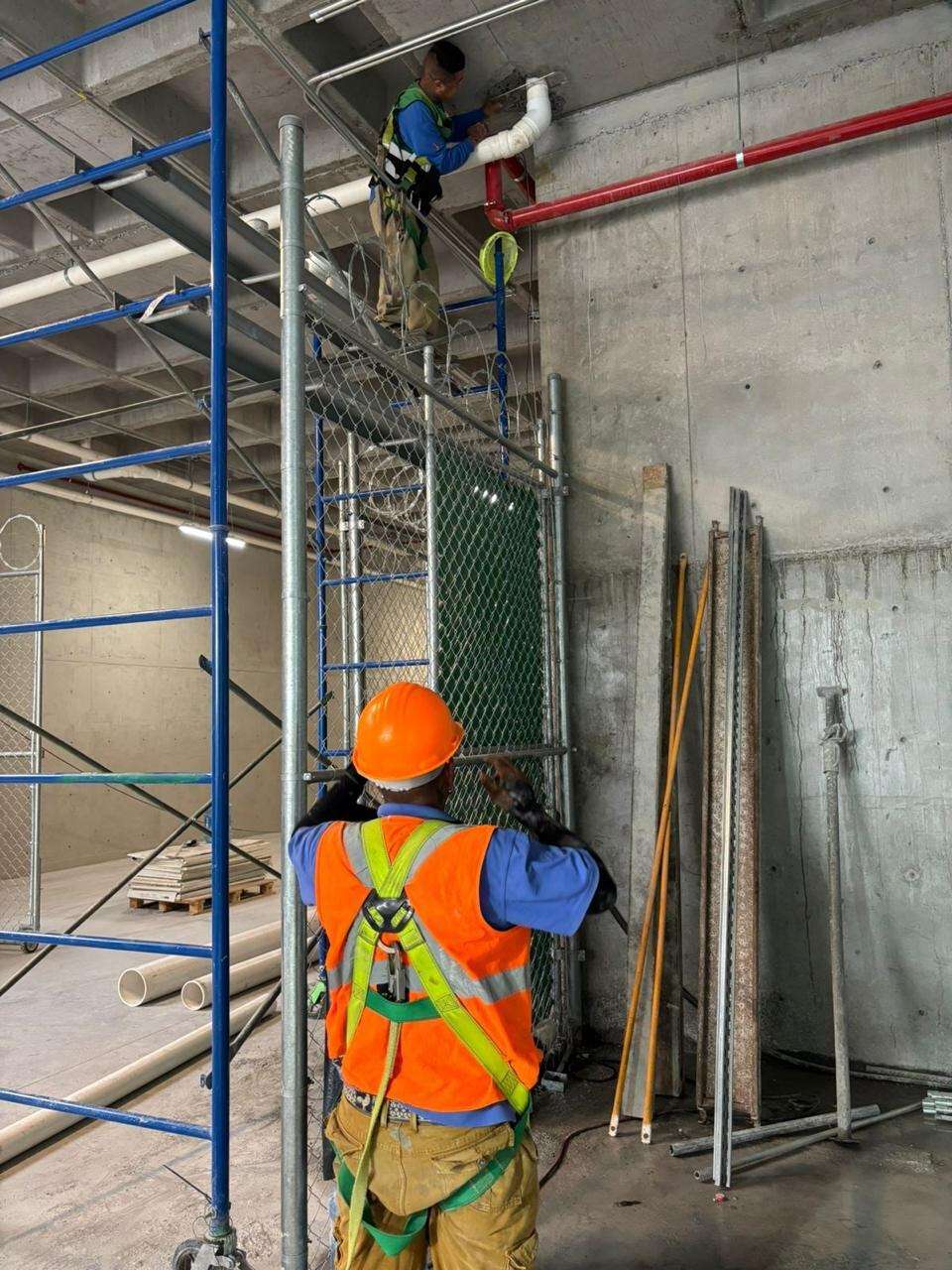 Trabajador con casco naranja, chaleco reflejante y arnés de cuerpo completo con línea de vida, montando tubería sobre un andamio, y otro abajo con el mismo equipo.