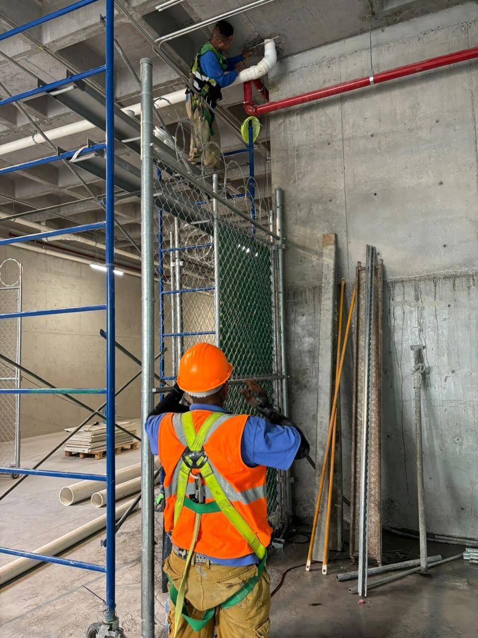 Trabajador con casco naranja, chaleco reflejante y arnés de cuerpo completo con línea de vida, montando tubería sobre un andamio, y otro abajo con el mismo equipo.