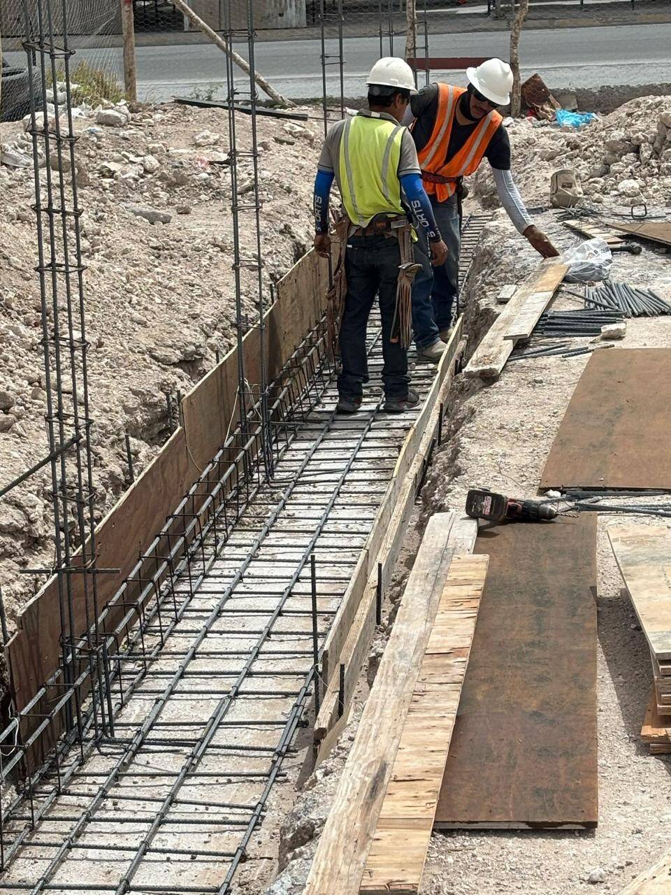 Dos trabajadores con casco blanco y chaleco parados sobre la cimbra de una zanja, revisando el armado de varilla de una trabe de cimentación al borde de la calle.