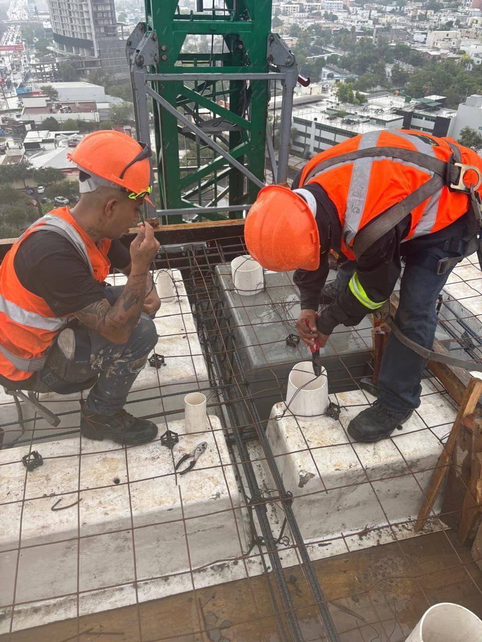 Dos trabajadores con casco naranja y chaleco reflejante habilitando el armado de acero de una losa, con la ciudad y una grúa torre al fondo.