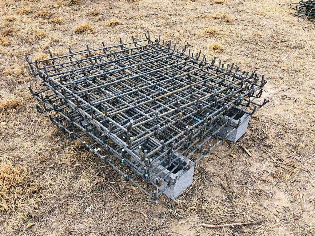 Parrilla de acero de refuerzo habilitada y apoyada sobre calzas de block en el terreno, con la maquinaria de la obra al fondo.