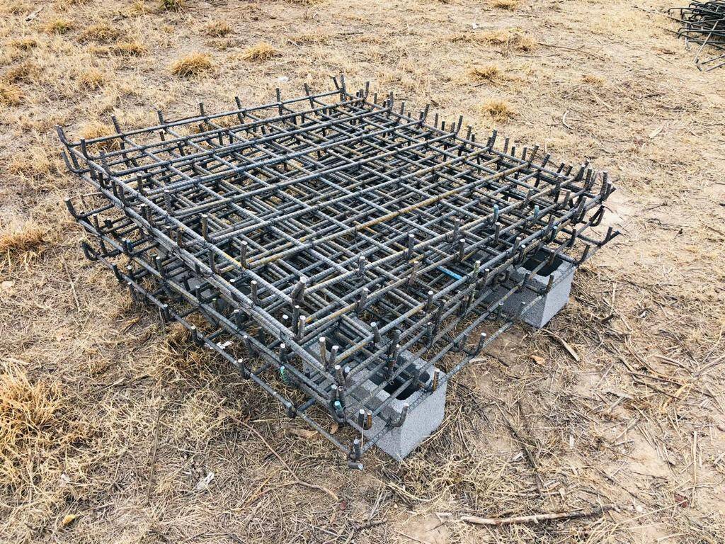 Parrilla de acero de refuerzo habilitada y apoyada sobre calzas de block en el terreno, con la maquinaria de la obra al fondo.