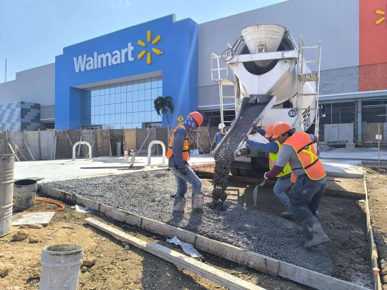Cuadrilla con casco y chaleco reflejante colando y emparejando una banqueta de concreto junto a una revolvedora, frente a la fachada azul de una tienda de autoservicio.