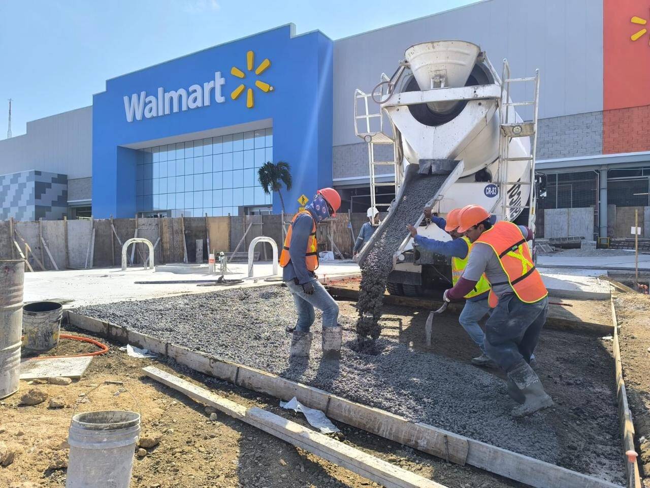 Cuadrilla con casco y chaleco reflejante colando y emparejando una banqueta de concreto junto a una revolvedora, frente a la fachada azul de una tienda de autoservicio.