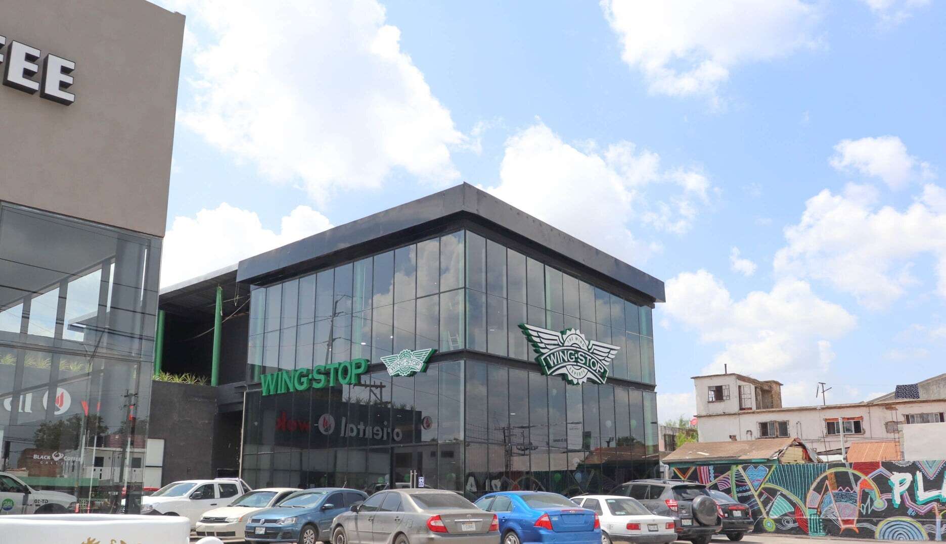 Edificio comercial de dos niveles con fachada de cristal y estructura oscura, visto desde el estacionamiento lleno de autos en un día despejado.