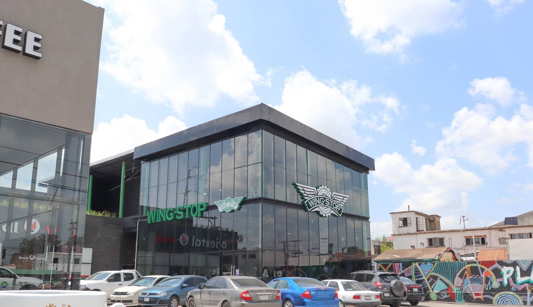 Edificio comercial de dos niveles con fachada de cristal y estructura oscura, visto desde el estacionamiento lleno de autos en un día despejado.