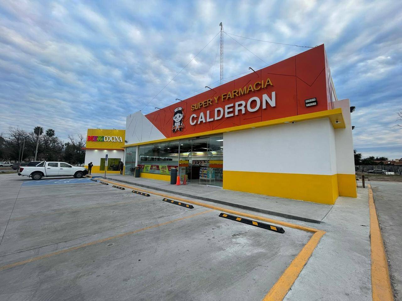 Fachada terminada de una Súper Farmacia Calderón vista en escorzo, con marquesina roja y amarilla, zócalo amarillo y el estacionamiento recién trazado bajo un cielo de nubes altas.