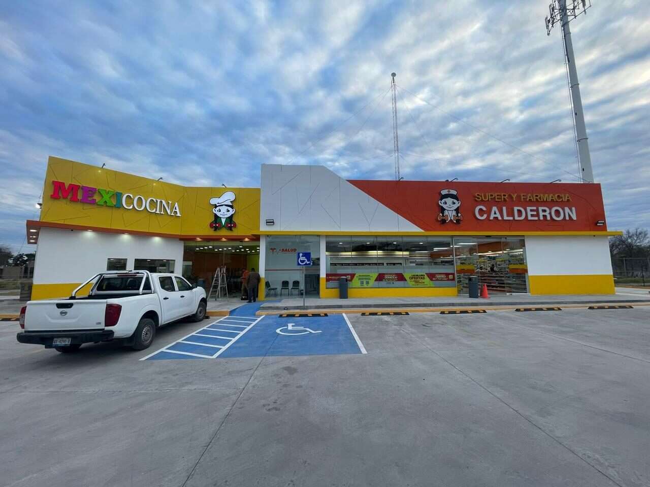 Fachada terminada de una Súper Farmacia Calderón con Mexicocina al lado, marquesina roja y blanca, zócalo amarillo y el estacionamiento trazado con cajón para persona con discapacidad.