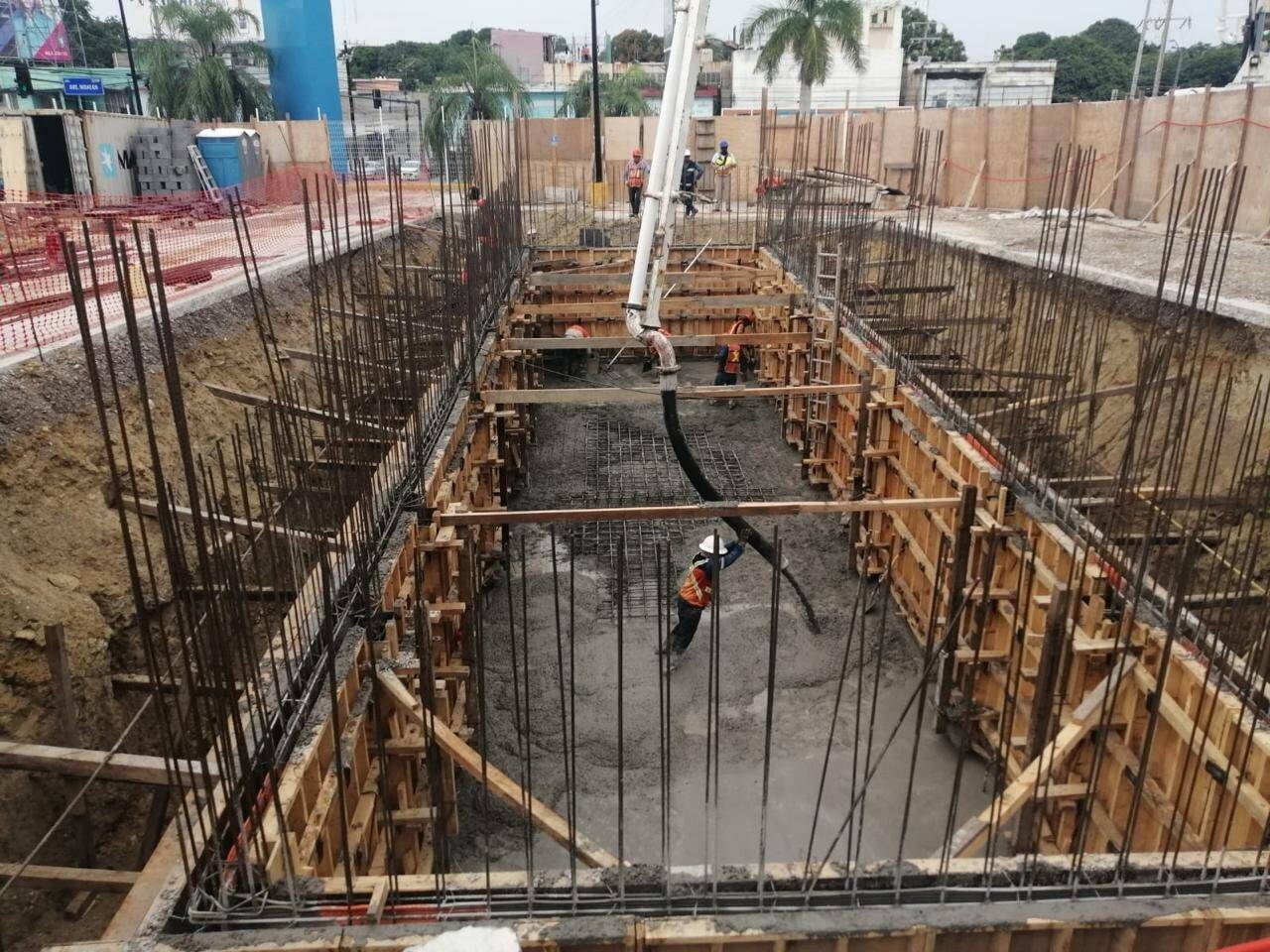 Excavación profunda de cimentación con los muros cimbrados en madera y el armado de acero saliendo del fondo, con la pluma de una bomba de concreto entrando al cajón y un trabajador con chaleco naranja adentro.