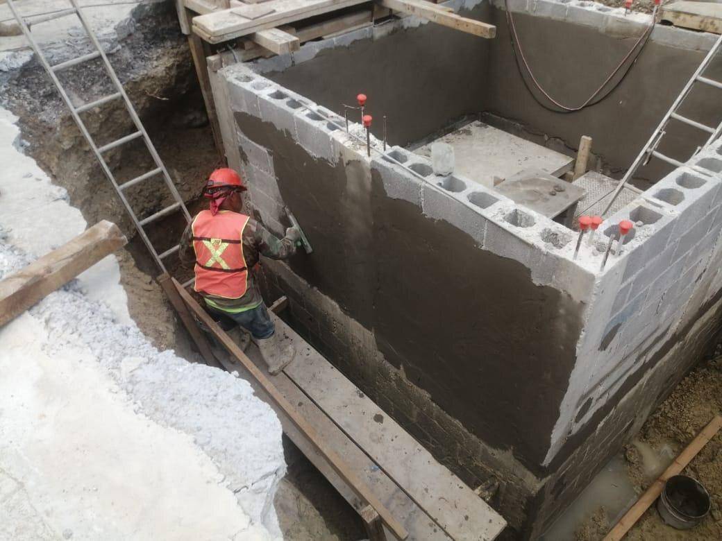 Trabajador con casco rojo y chaleco naranja dentro de una cisterna de muros de block ya aplanados, con la escalera de acceso apoyada en el borde.