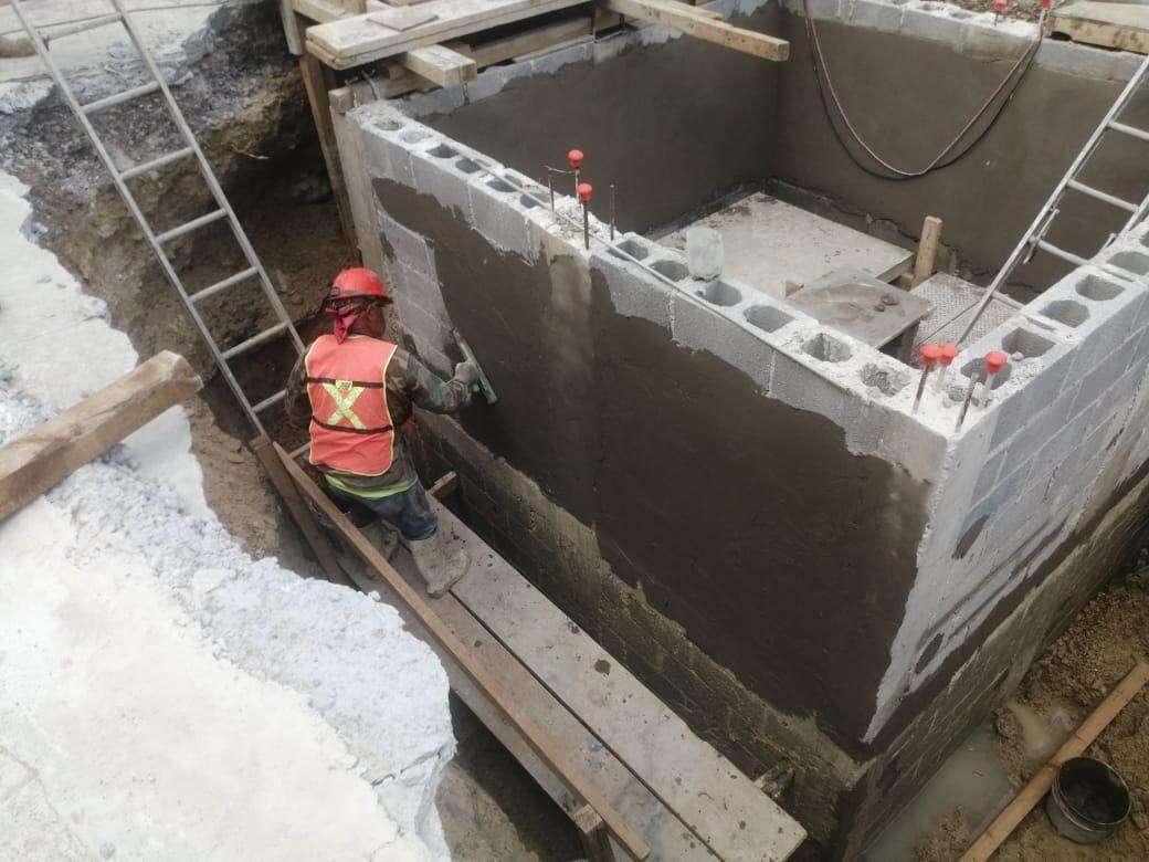 Trabajador con casco rojo y chaleco naranja dentro de una cisterna de muros de block ya aplanados, con la escalera de acceso apoyada en el borde.