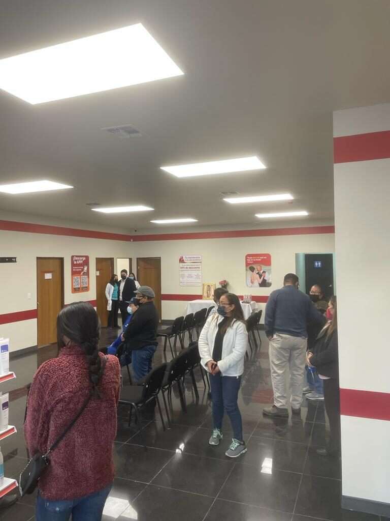 Sala de espera de una clínica el día de su apertura, con personas formadas frente al mostrador, sillas negras alineadas contra el muro y piso de loseta oscura brillante.