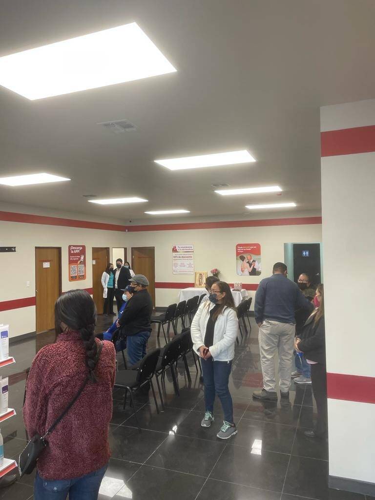 Sala de espera de una clínica el día de su apertura, con personas formadas frente al mostrador, sillas negras alineadas contra el muro y piso de loseta oscura brillante.
