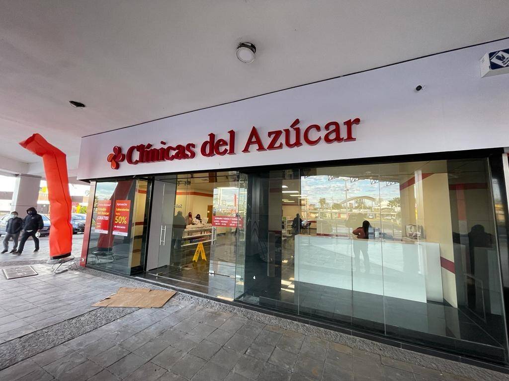 Fachada de una sucursal de Clínicas del Azúcar recién entregada, con cancelería de cristal de piso a techo, letrero rojo iluminado y un muñeco inflable naranja en la banqueta.