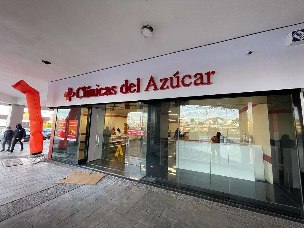 Fachada de una sucursal de Clínicas del Azúcar recién entregada, con cancelería de cristal de piso a techo, letrero rojo iluminado y un muñeco inflable naranja en la banqueta.