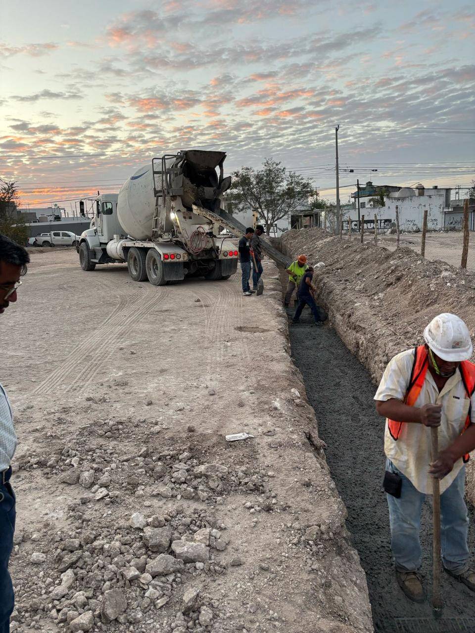 Revolvedora descargando concreto en una zanja larga al atardecer, con un trabajador de casco blanco y chaleco naranja emparejando la mezcla con una pala.