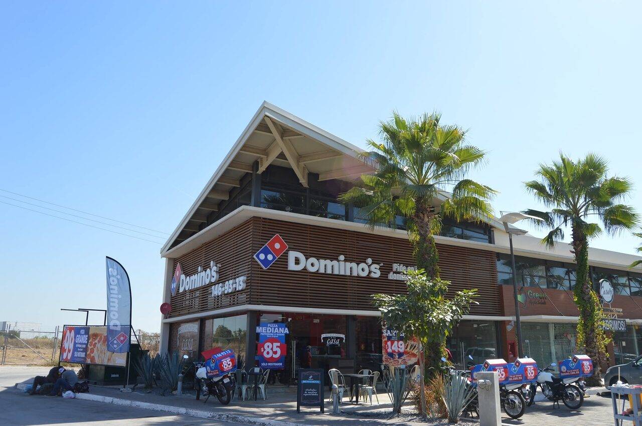 Edificio de dos niveles con una sucursal de Domino’s en planta baja, con palmeras al frente y motocicletas de reparto estacionadas.