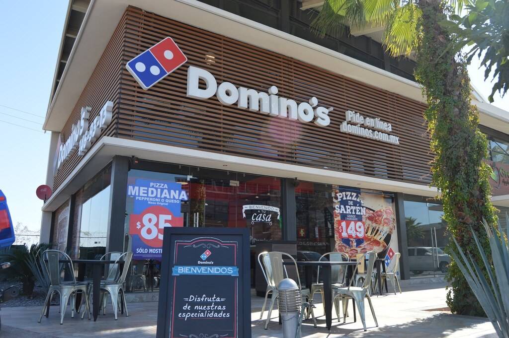 Fachada de una pizzería Domino’s terminada, con celosía de madera sobre el acceso, franja azul con las promociones y palmas sembradas al frente.