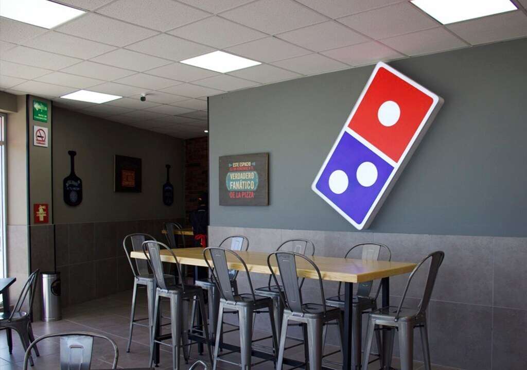 Interior terminado de una pizzería con muro gris, el logotipo de Domino’s en relieve, un paño de tabique aparente y mesas de madera con bancos metálicos grises.