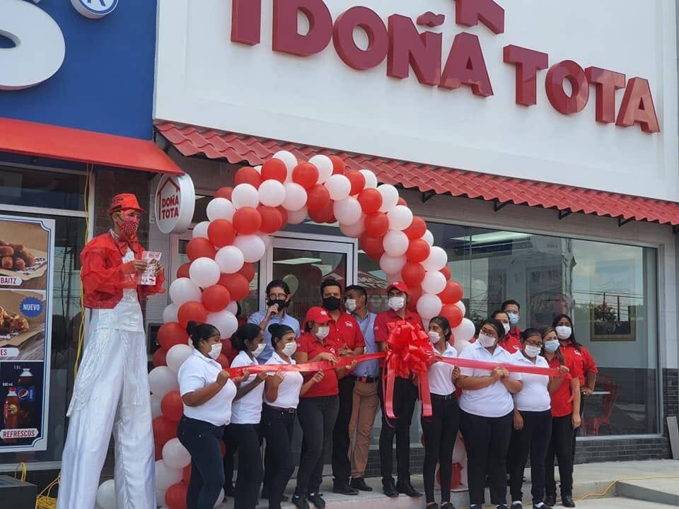 Corte de listón en la entrada de una sucursal de Doña Tota, con el personal formado bajo un arco de globos rojos y blancos y una figura de zancos a un costado.