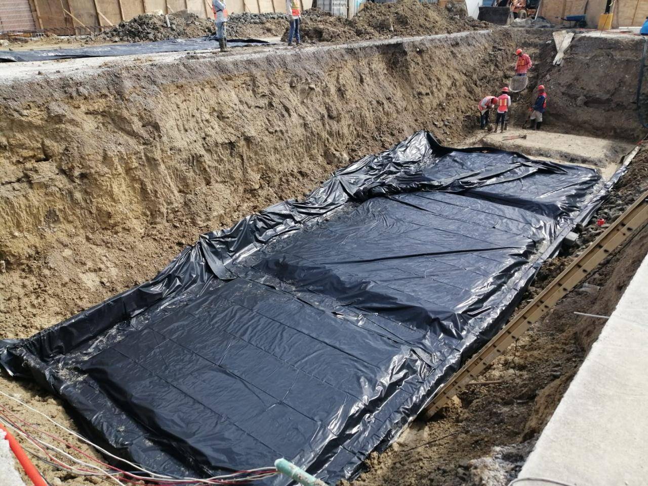 Fosa rectangular excavada y forrada con membrana negra, con el brocal de concreto colado alrededor y la barda de la obra al fondo.