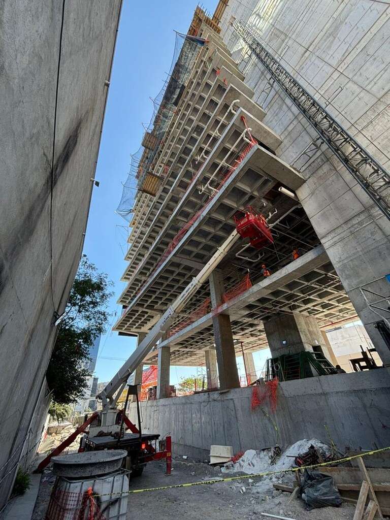 Edificio en obra visto desde abajo, con las losas reticulares de concreto en voladizo, malla de protección en los niveles altos y una plataforma roja de maniobra colgada del borde.