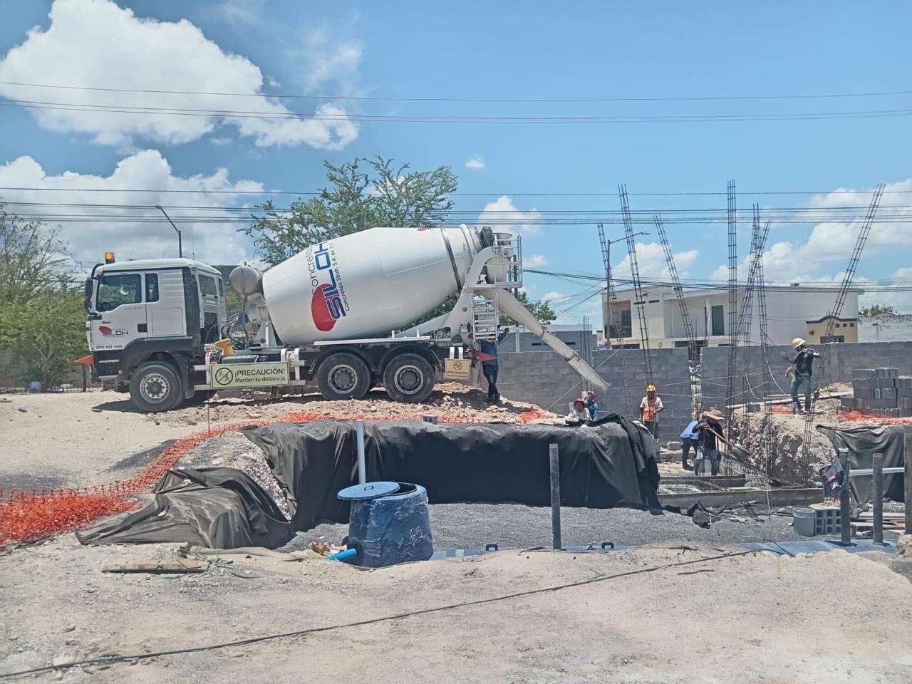 Revolvedora blanca descargando concreto en la obra de una estación de servicio, junto a una excavación forrada con membrana negra, con torres de alta tensión al fondo.