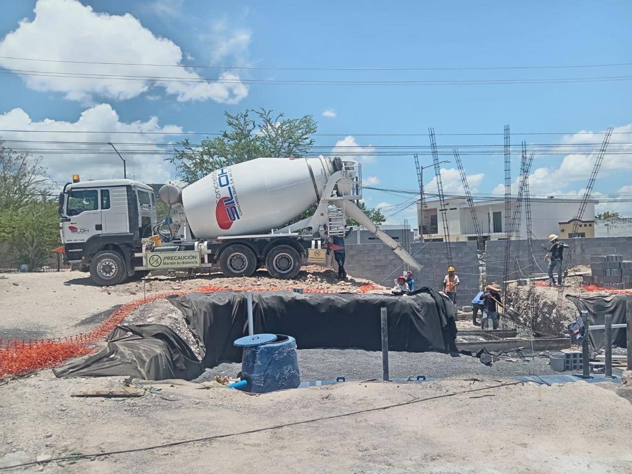 Revolvedora blanca descargando concreto en la obra de una estación de servicio, junto a una excavación forrada con membrana negra, con torres de alta tensión al fondo.