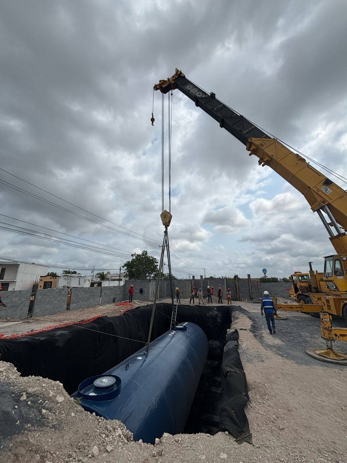 Grúa amarilla bajando un tanque cilíndrico azul a una fosa forrada con membrana negra, bajo un cielo nublado, con la cuadrilla de casco a distancia y el muro de block al fondo.