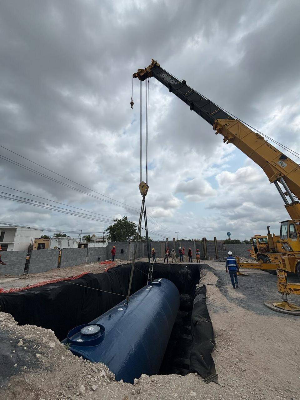 Grúa amarilla bajando un tanque cilíndrico azul a una fosa forrada con membrana negra, bajo un cielo nublado, con la cuadrilla de casco a distancia y el muro de block al fondo.
