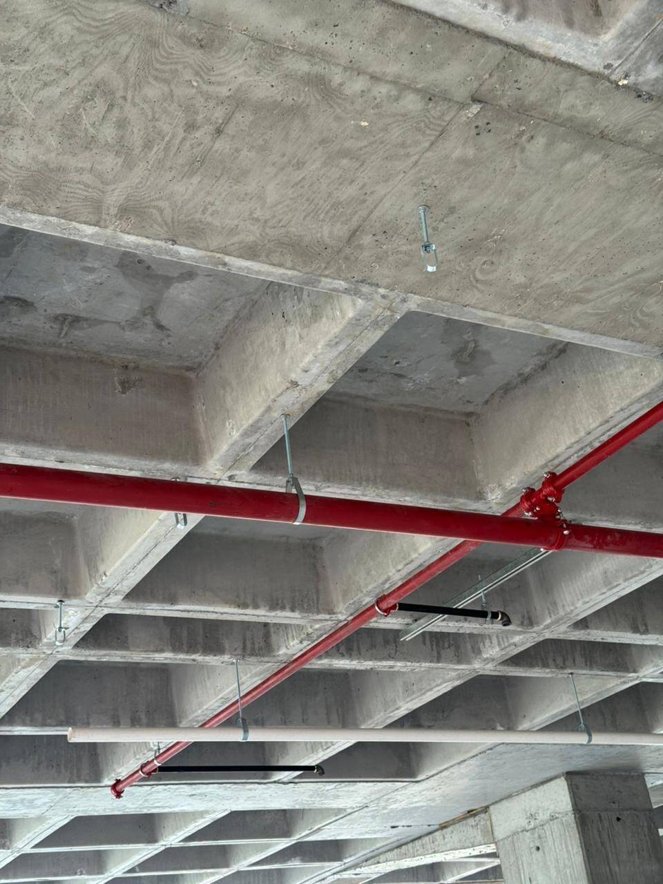 Losa reticular de concreto aparente vista desde abajo, con la tubería roja de la red contra incendio y los ramales sanitarios de PVC blanco colgados de ella.