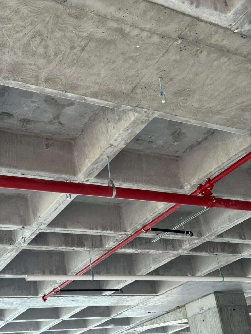 Losa reticular de concreto aparente vista desde abajo, con la tubería roja de la red contra incendio y los ramales sanitarios de PVC blanco colgados de ella.