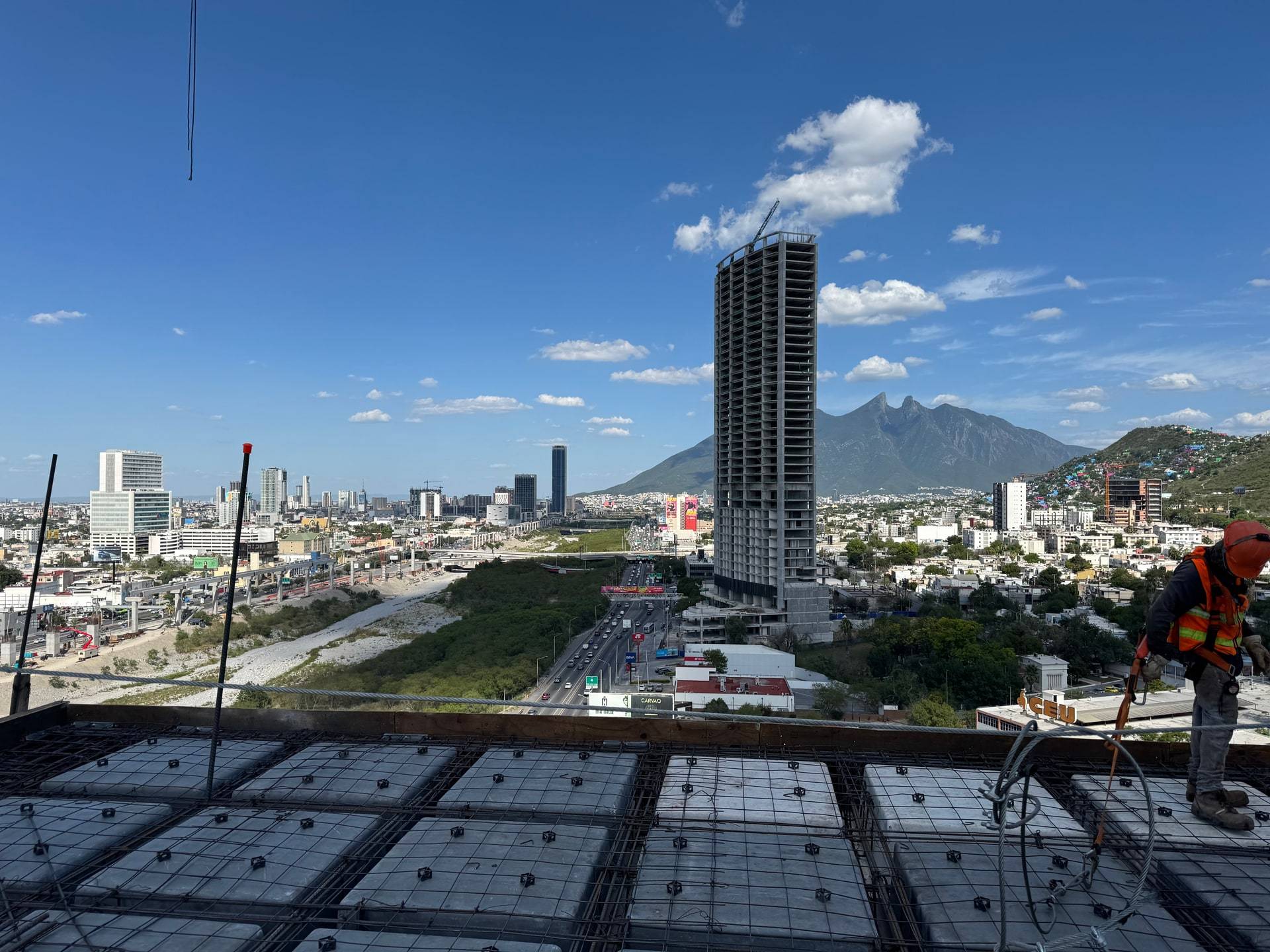 Losa de un edificio en obra con el armado y los casetones ya colocados, vista hacia la ciudad: al frente el nivel en proceso, al fondo una torre en obra gris y el Cerro de la Silla, y a la derecha un trabajador con casco naranja, chaleco reflejante y arnés parado en el borde.