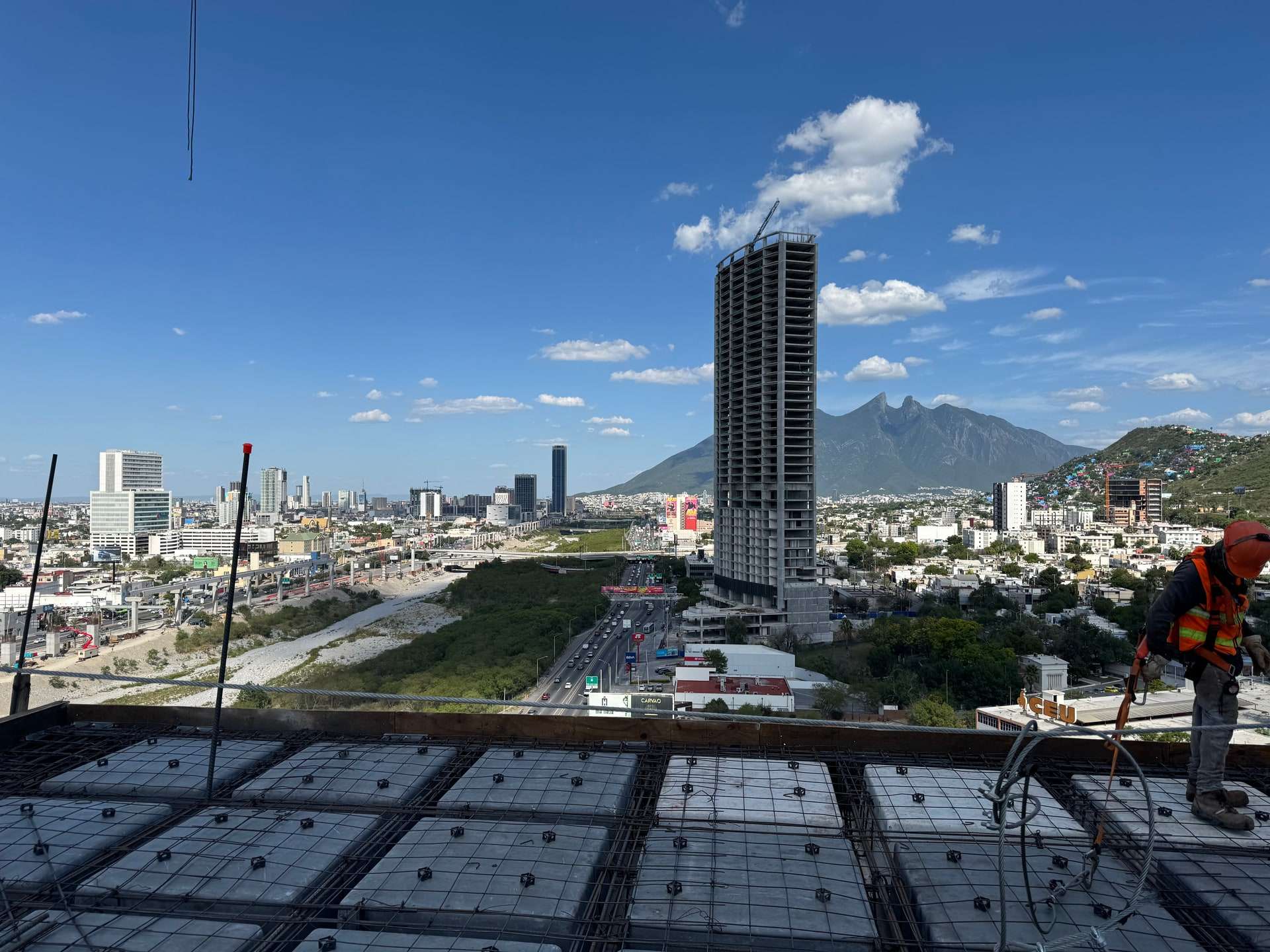 Losa de un edificio en obra con el armado y los casetones ya colocados, vista hacia la ciudad: al frente el nivel en proceso, al fondo una torre en obra gris y el Cerro de la Silla, y a la derecha un trabajador con casco naranja, chaleco reflejante y arnés parado en el borde.