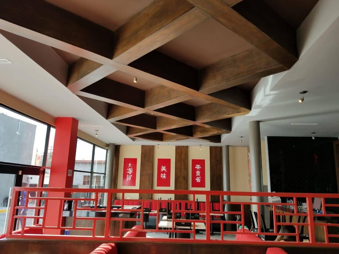 Entrepiso terminado de un restaurante, con plafón artesonado de madera, barandal de herrería roja y las mesas ya colocadas junto al ventanal.