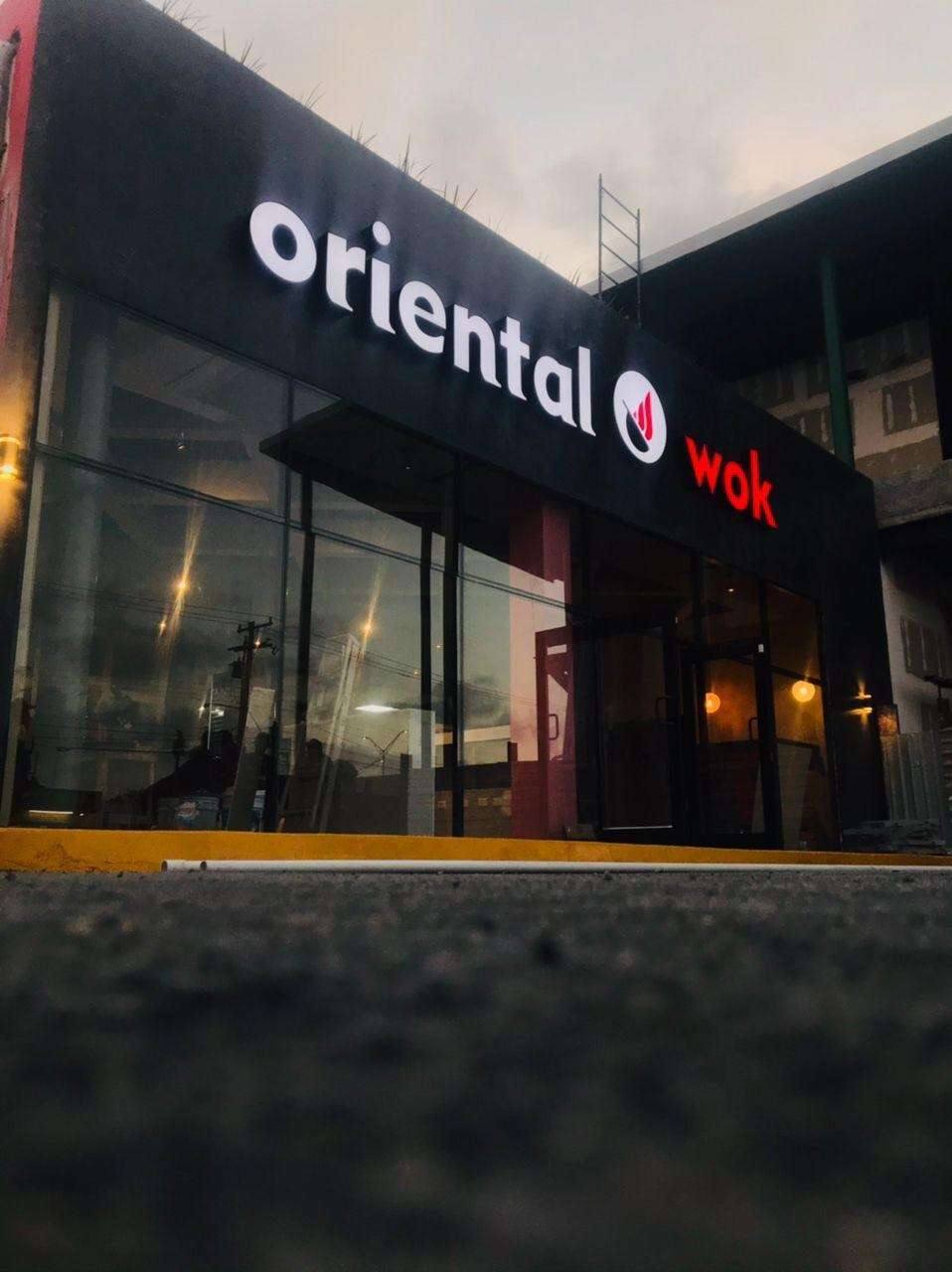 Fachada de un restaurante Oriental Wok al anochecer, con el frente negro, cancelería de piso a techo y las letras blancas y rojas del anuncio encendidas.