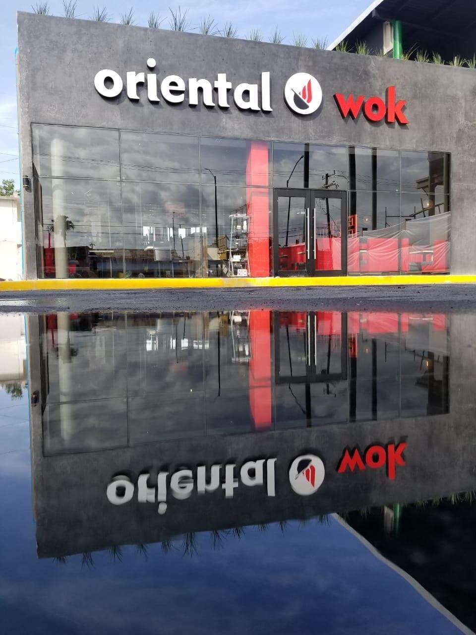 Fachada de Oriental Wok reflejada en un charco del estacionamiento, con el anuncio y la cancelería duplicados en el agua.