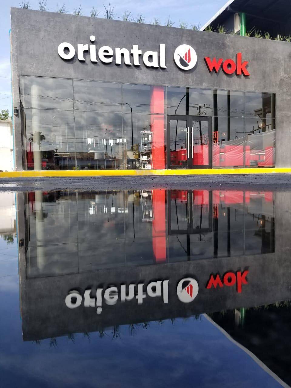 Fachada de Oriental Wok reflejada en un charco del estacionamiento, con el anuncio y la cancelería duplicados en el agua.