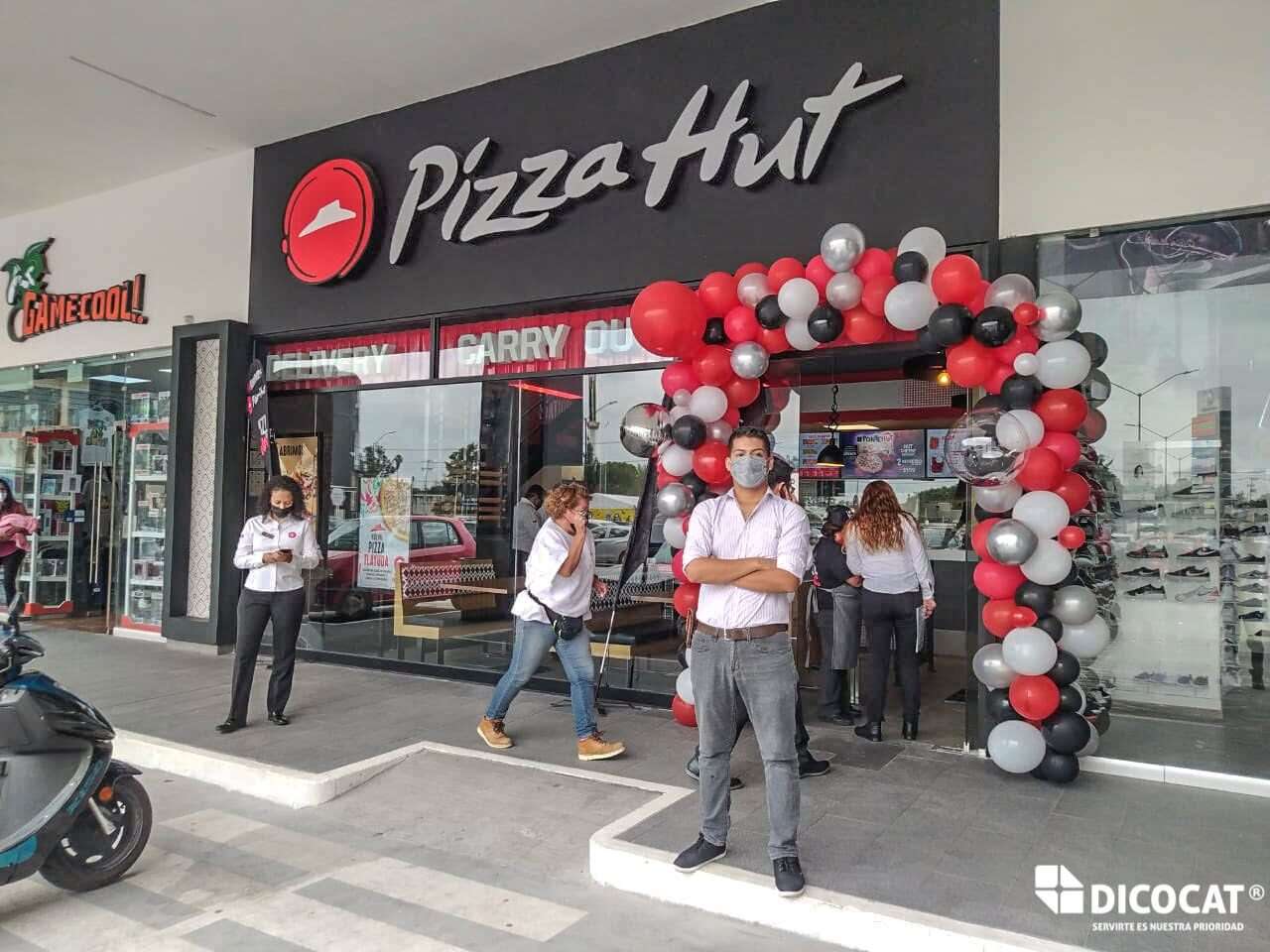 Fachada de una sucursal de Pizza Hut en plaza comercial, con arco de globos rojos, grises y blancos y el personal en la banqueta.