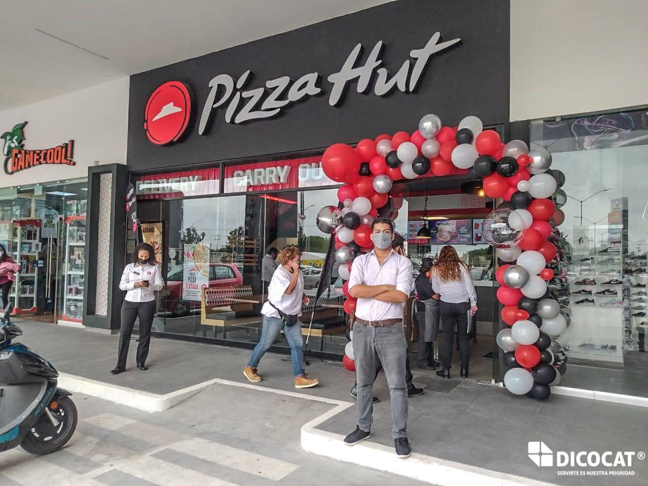 Fachada de una sucursal de Pizza Hut en plaza comercial, con arco de globos rojos, grises y blancos y el personal en la banqueta.