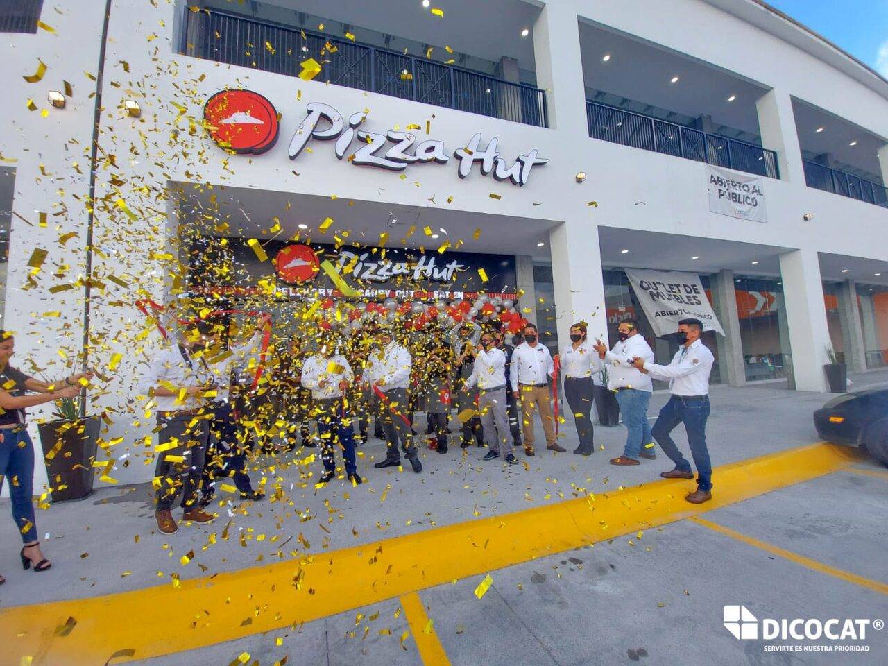 Fachada de una sucursal de Pizza Hut en el momento de la apertura, con una lluvia de confeti dorado cayendo sobre el personal reunido en la banqueta.