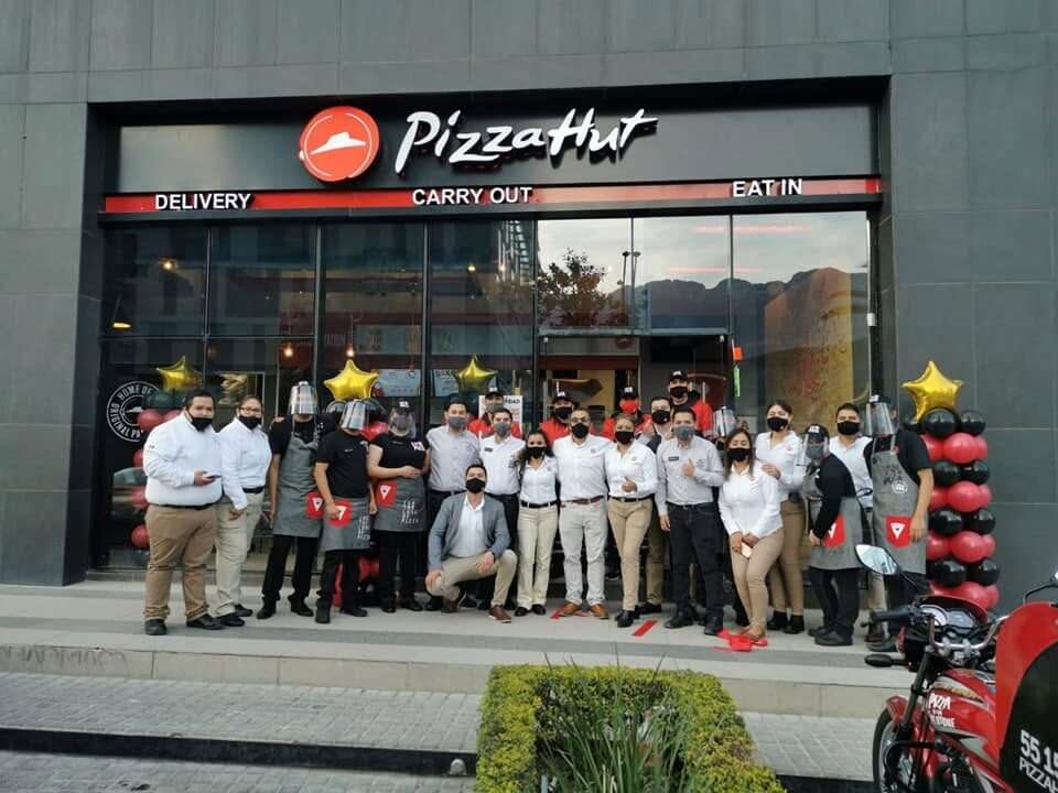 Personal de una sucursal de Pizza Hut formado frente a la fachada terminada el día de la apertura, con el listón puesto y motocicletas de reparto al frente.