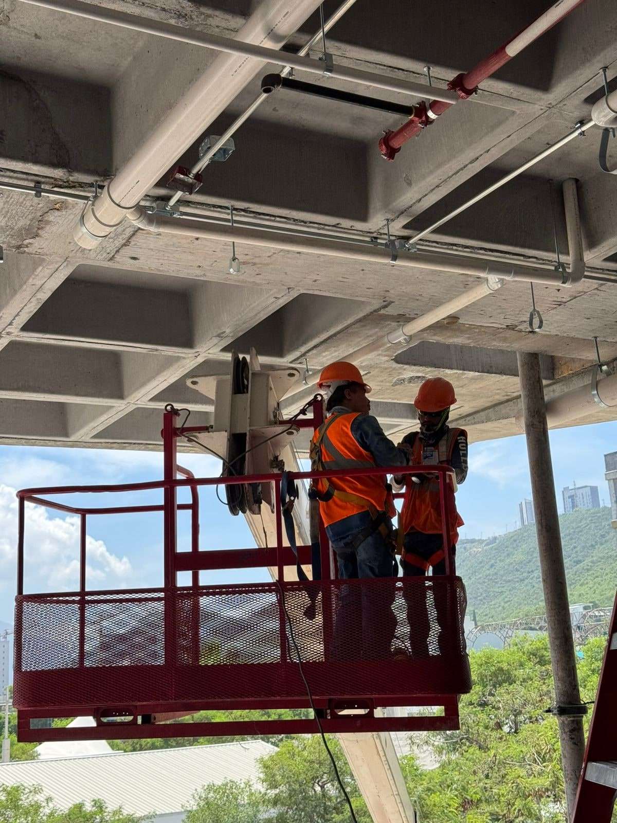 Dos trabajadores con casco naranja, chaleco reflejante y arnés montando desde una canastilla elevadora roja la tubería de la red contra incendio bajo una losa reticular de concreto aparente, con el cerro y la ciudad al fondo.