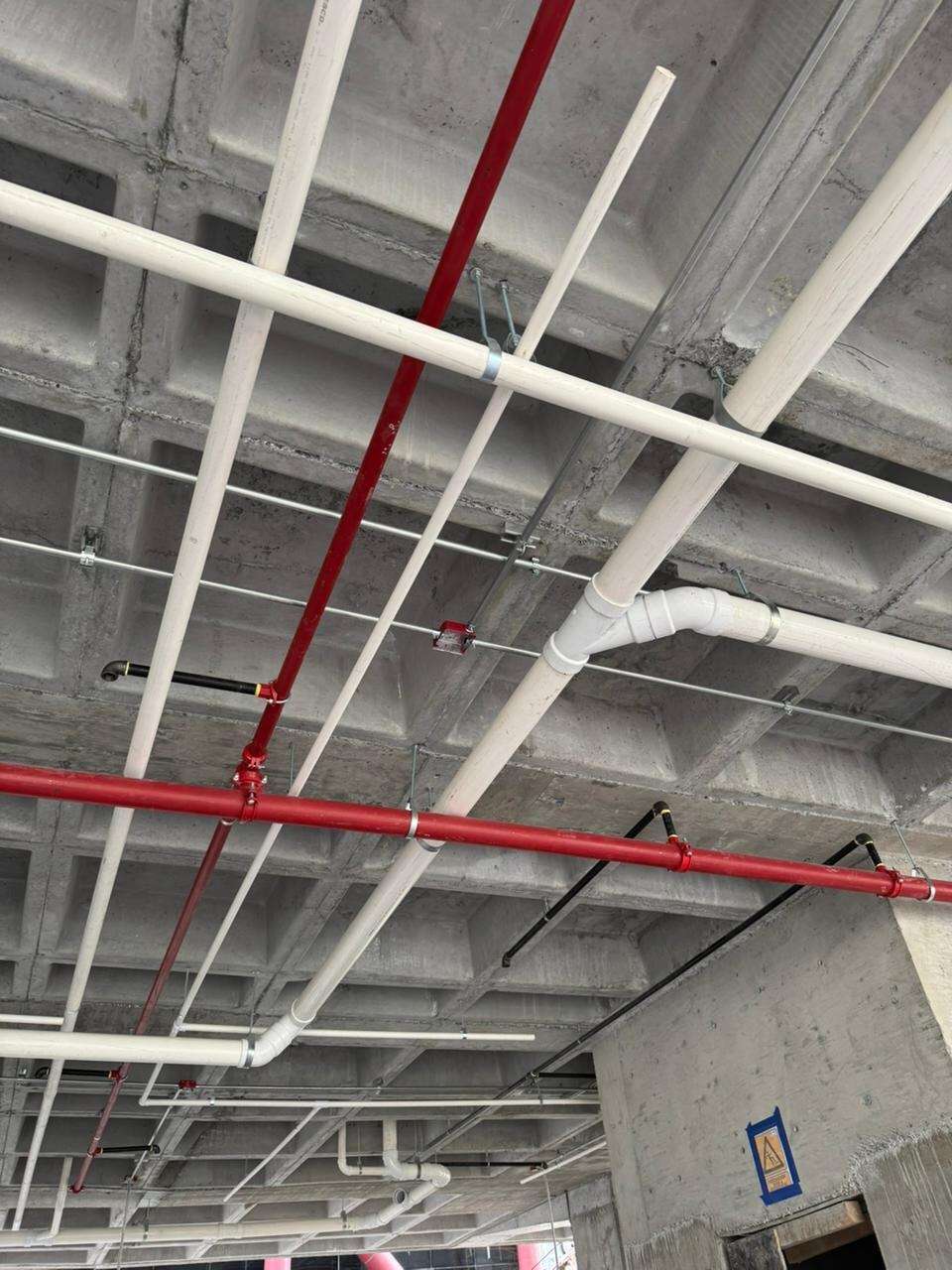 Tubería de red contra incendio pintada de rojo y ramales sanitarios de PVC blanco colgados bajo una losa reticular de concreto aparente.