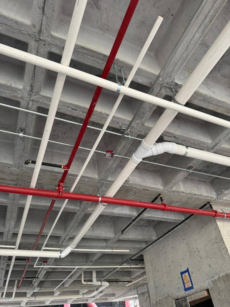 Tubería de red contra incendio pintada de rojo y ramales sanitarios de PVC blanco colgados bajo una losa reticular de concreto aparente.