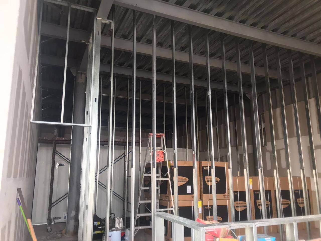 Interior de un local en obra con la estructura metálica de los muros de tablarroca montada y la cubierta de lámina a la vista, con una escalera de tijera al fondo.