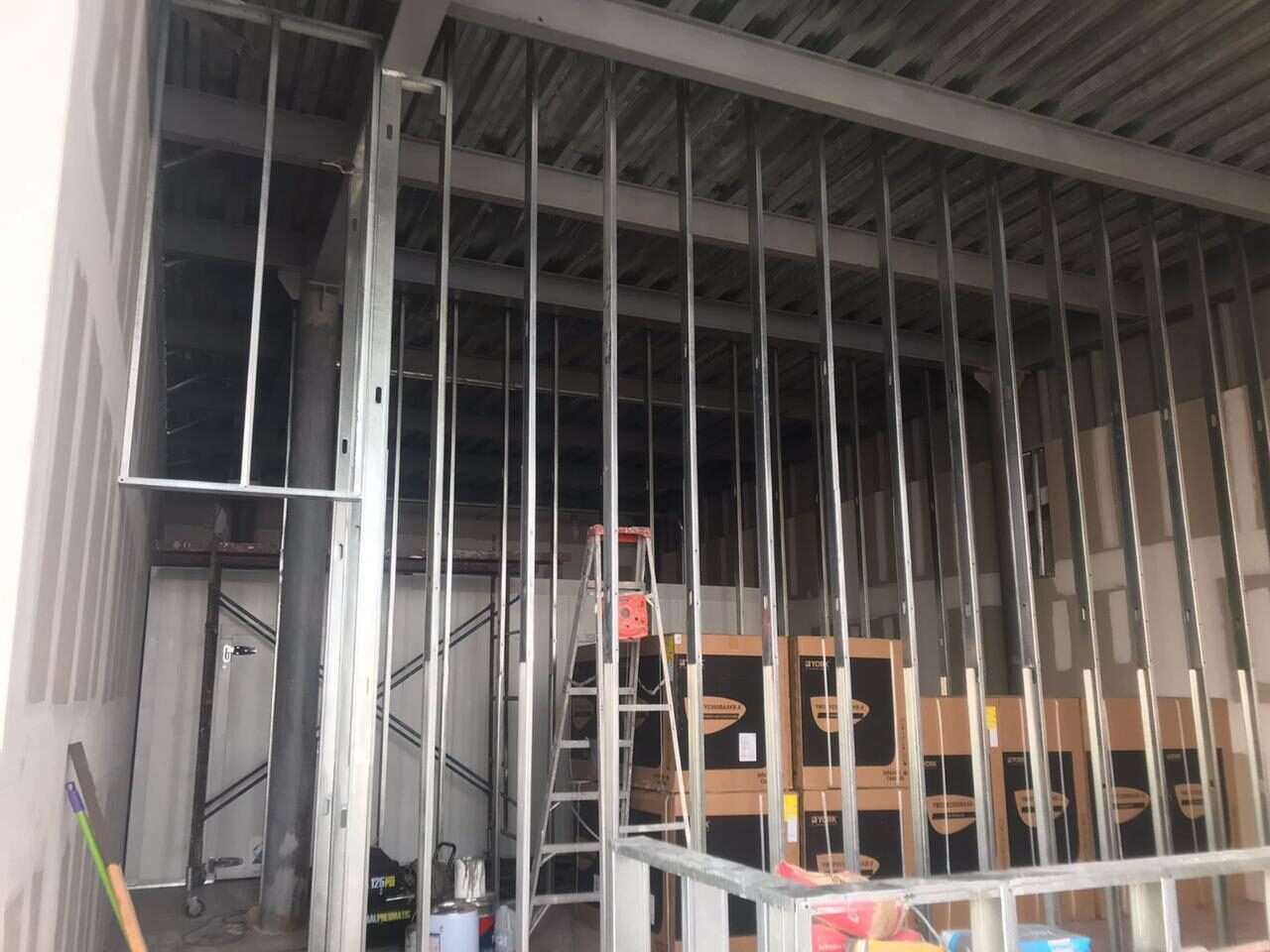 Interior de un local en obra con la estructura metálica de los muros de tablarroca montada y la cubierta de lámina a la vista, con una escalera de tijera al fondo.