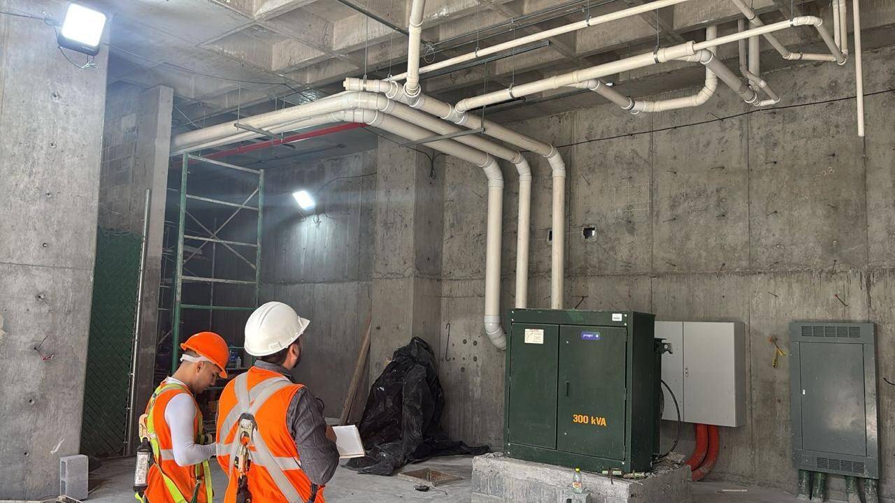 Dos personas con casco blanco y chaleco naranja revisando un tablero eléctrico verde montado en un local en obra, con la canalización y los ductos a la vista bajo la losa.
