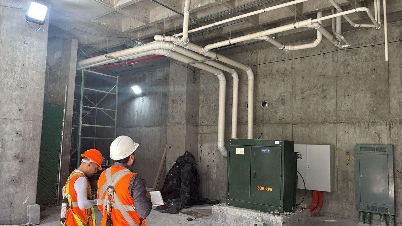Dos personas con casco blanco y chaleco naranja revisando un tablero eléctrico verde montado en un local en obra, con la canalización y los ductos a la vista bajo la losa.