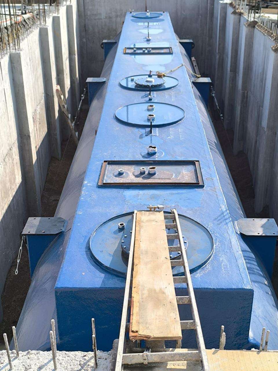 Fosa excavada y forrada con membrana negra con dos tanques cilíndricos azules ya colocados en el fondo, vista desde el borde.